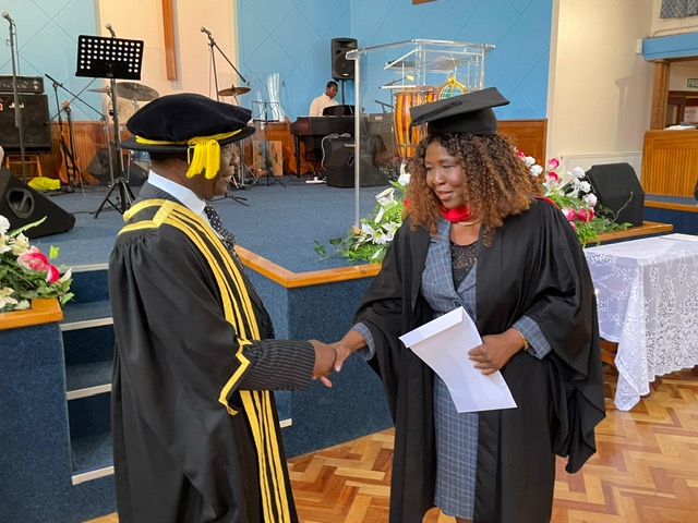 Two people in academic robes shaking hands beside a stage